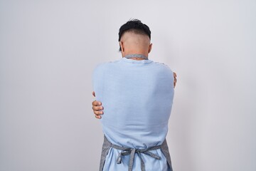 Hispanic young man wearing apron over white background hugging oneself happy and positive from...