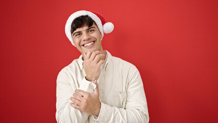 Naklejka premium Young hispanic man smiling confident wearing christmas hat over isolated red background