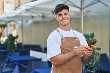 Young hispanic man waiter smiling confident using smartphone at coffee shop terrace
