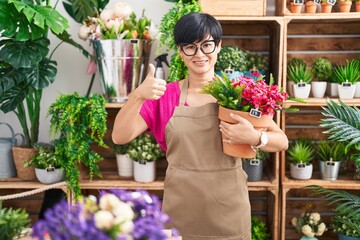 Young asian woman with short hair working at florist shop holding plant smiling happy and positive, thumb up doing excellent and approval sign