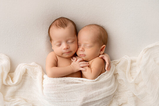 Tiny newborn twins boys in white cocoons on a white background. A newborn twin sleeps next to his brother. Newborn two twins boys hugging each other. Professional studio photography. 