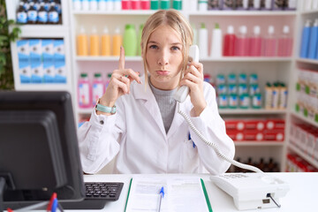 Young caucasian woman working at pharmacy drugstore speaking on the telephone pointing up looking...
