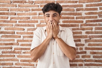 Arab man with beard standing over bricks wall background laughing and embarrassed giggle covering...