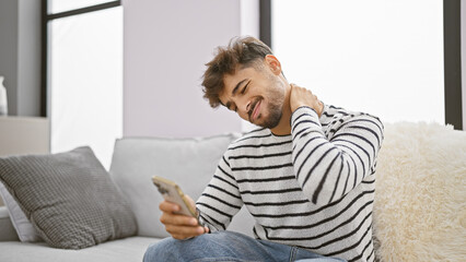 Exhausted young arabian man at home, using smartphone on sofa, suffering serious cervical pain, stressed from texting