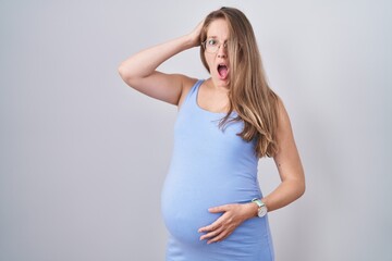 Young pregnant woman standing over white background crazy and scared with hands on head, afraid and...