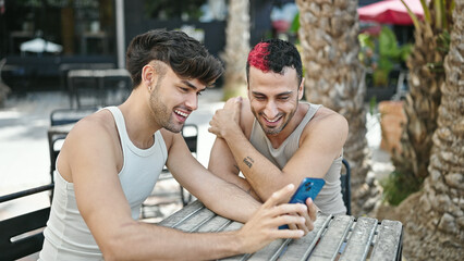 Two men couple smiling confident using smartphone at coffee shop terrace