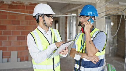 Two men builders talking on smartphone reading document at construction site