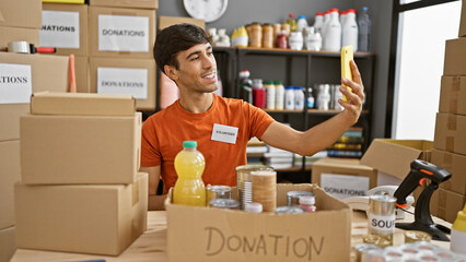 Smiling young hispanic man volunteer taking an altruistic selfie with a cellphone in a cheerful charity center, making memories while working indoors
