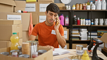 Young hispanic man volunteer talking on smartphone taking notes at charity center