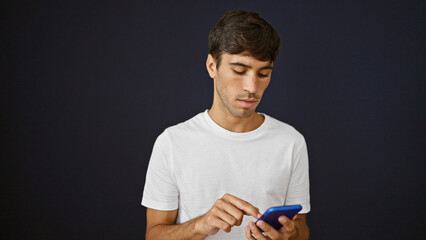 Cool young hispanic man engrossed in avidly texting a serious message online. he uses smartphone technology with concentrated expression, isolated against black background wall, lifestyle portrait.