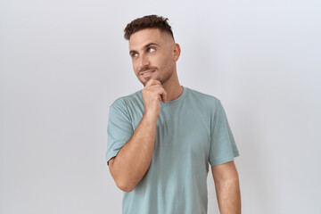 Hispanic man with beard standing over white background with hand on chin thinking about question, pensive expression. smiling and thoughtful face. doubt concept.