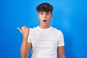 Hispanic teenager standing over blue background surprised pointing with hand finger to the side,...