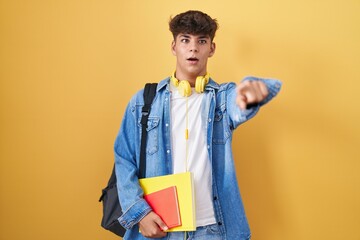 Hispanic teenager wearing student backpack and holding books pointing with finger surprised ahead,...