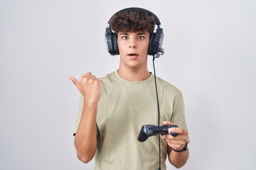 Hispanic teenager playing video game holding controller surprised pointing with hand finger to the side, open mouth amazed expression. © Krakenimages.com
