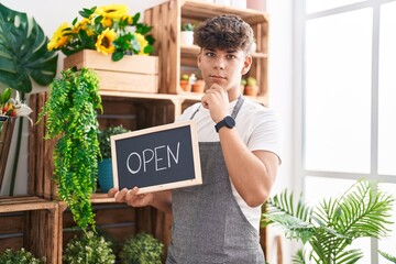 Hispanic teenager working at florist holding open sign serious face thinking about question with...