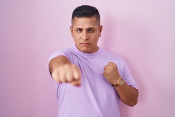 Young hispanic man standing over pink background punching fist to fight, aggressive and angry...