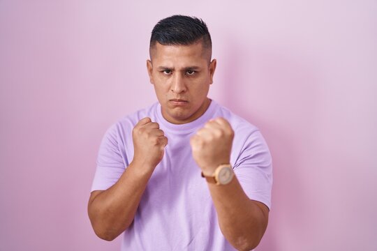 Young hispanic man standing over pink background ready to fight with fist defense gesture, angry and upset face, afraid of problem