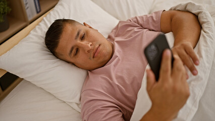 Young latin man using smartphone lying on bed at bedroom