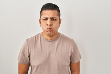 Hispanic young man standing over white background puffing cheeks with funny face. mouth inflated...