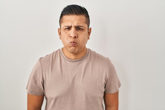 Hispanic young man standing over white background puffing cheeks with funny face. mouth inflated with air, crazy expression.