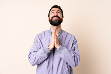 Caucasian man with beard wearing a jacket over isolated background keeps palm together. Person asks...