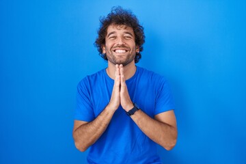 Hispanic young man standing over blue background praying with hands together asking for forgiveness...