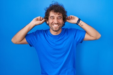 Hispanic young man standing over blue background smiling pulling ears with fingers, funny gesture....