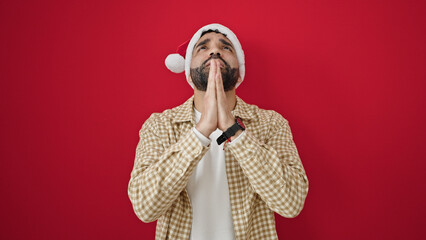 Young hispanic man praying wearing christmas hat over isolated red background
