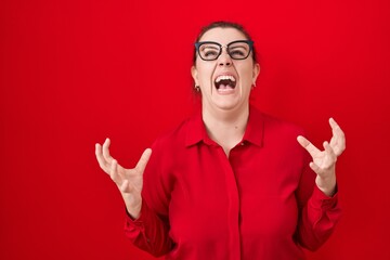 Naklejka premium Young hispanic woman with red hair standing over red background crazy and mad shouting and yelling with aggressive expression and arms raised. frustration concept.