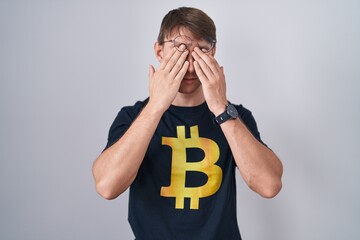 Caucasian blond man wearing bitcoin t shirt rubbing eyes for fatigue and headache, sleepy and tired...