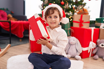Obraz na płótnie Canvas Adorable hispanic boy hearing gift sound sitting on floor by christmas tree at home