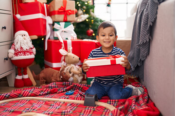 Adorable hispanic toddler holding gift sitting on floor by christmas tree at home