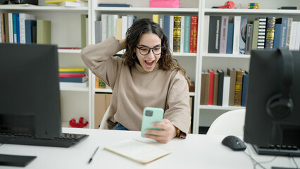 Young beautiful hispanic woman student using smartphone with surprise expression at library...