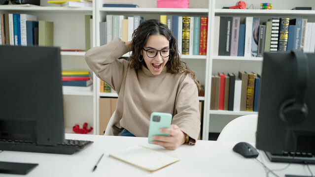 Young beautiful hispanic woman student using smartphone with surprise expression at library university