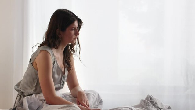 A young woman in a gray nightgown is sitting on the bed and thinking about something. A woman who woke up early in the morning sits on the bed and mentally prepares for a hard working day.