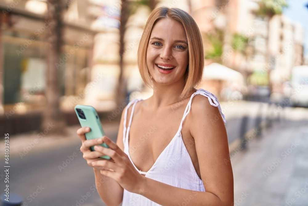 Wall mural Young blonde woman smiling confident using smartphone at street