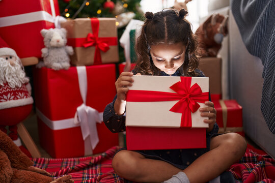Adorable hispanic girl unpacking gift sitting on floor by christmas tree at home