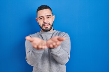 Hispanic man standing over blue background smiling with hands palms together receiving or giving...