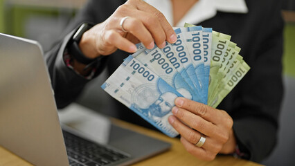 Young beautiful hispanic woman business worker holding chilean pesos using laptop at office