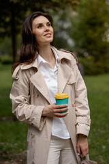 A charming young girl relaxes in a spring park outdoors with a cup of coffee after study or work. A smiling positive lady model within a pensive look is posing in full height for snapshots of