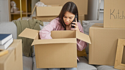 Young beautiful hispanic woman speaking on the phone unpacking cardboard box at new home