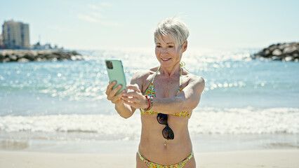 Middle age blonde woman tourist wearing bikini using smartphone at the beach