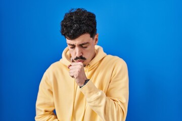 Hispanic man standing over blue background feeling unwell and coughing as symptom for cold or...
