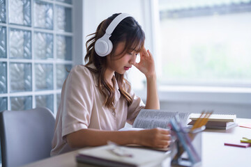 Young asian student in casual clothes wearing headphones to listening the music and reading textbook to thinking ideas while studying education knowledge and doing homework assignments in university - obrazy, fototapety, plakaty