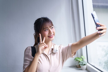 Young asian student in casual clothes showing v sign gesture with happiness and selfie on smartphone to relaxation after reading book and studying education knowledge to doing homework in university