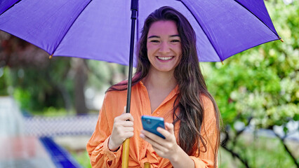 Young beautiful hispanic woman holding umbrella using smartphone at park