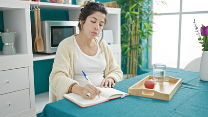 Young pregnant woman having breakfast writing on notebook at dinning room
