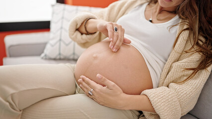 Young pregnant woman sitting on sofa applying lotion on belly at home