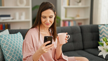 Young beautiful hispanic woman using smartphone drinking coffee at home
