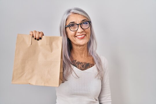 Middle age woman with grey hair holding take away paper bag looking positive and happy standing and smiling with a confident smile showing teeth